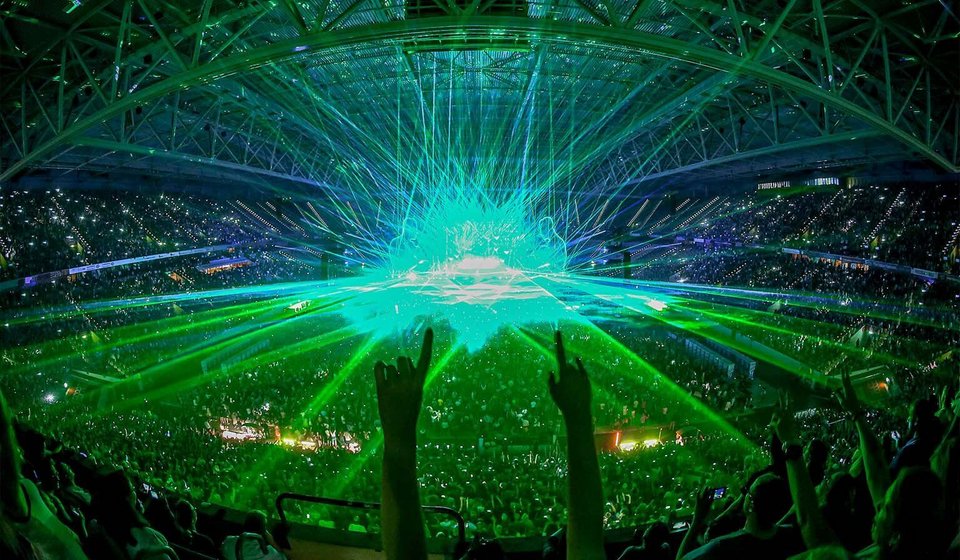 Konzert in der Arena bei geschlossenem Dach. Von der Bühne strahlen grüne und blaue Lichter die Halle an. Zentral werden Zwei Hände in die Luft gehalten. 