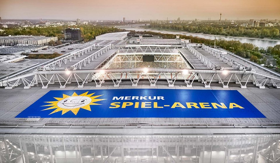Drohnenaufnahme MERKUR SPIEL-ARENA von außen. Auf dem Dach sieht man das Logo der Arena. Im Hintergrund fließt der Rhein und man sieht den Rheinturm vor dem orangenen Himmel. 
